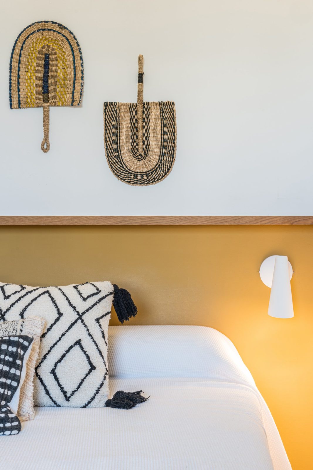 Bed against an ochre painted headboard band with woven fans and a white wall light