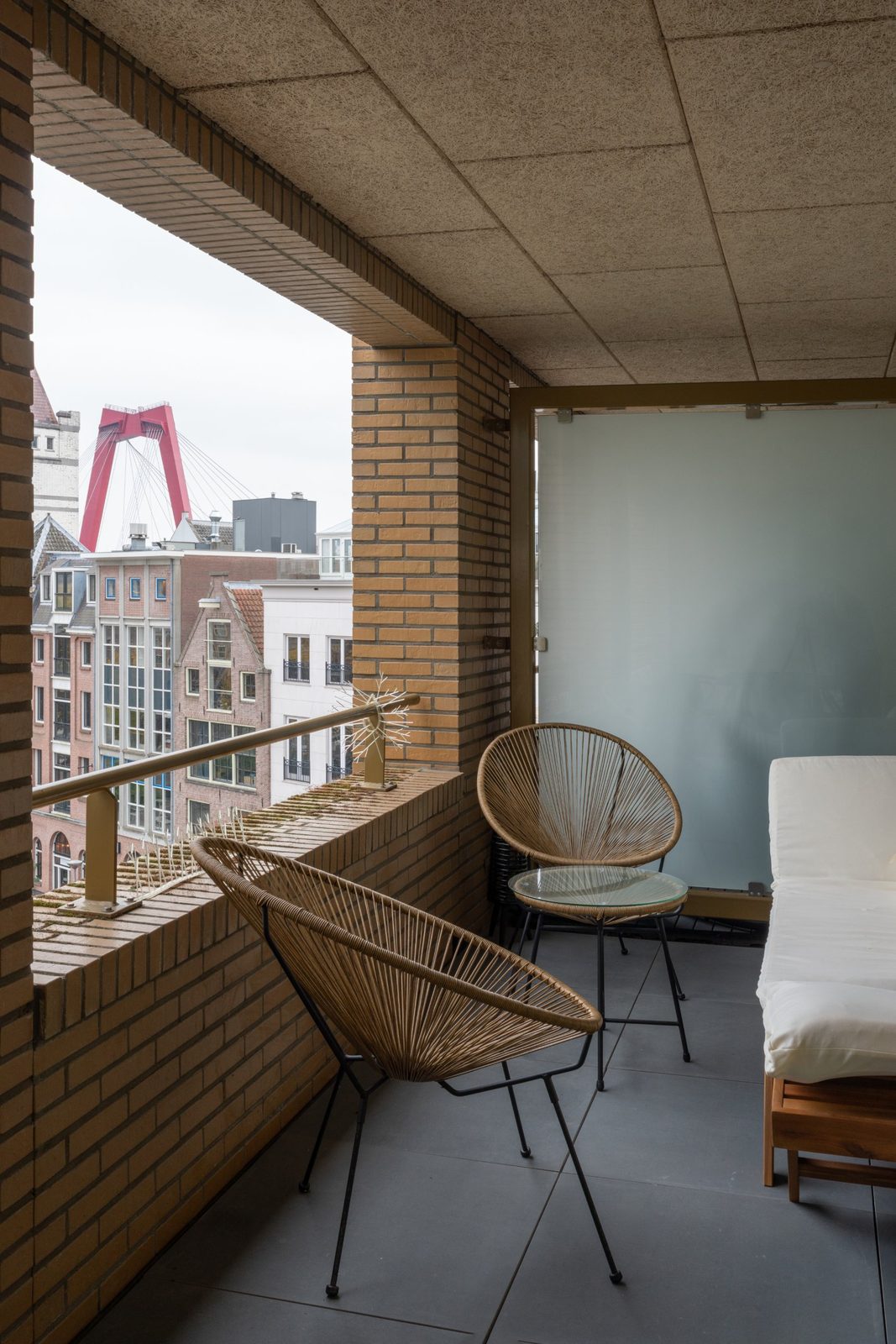 Covered brick balcony with woven chairs and a view of the Willemsbrug bridge in Rotterdam