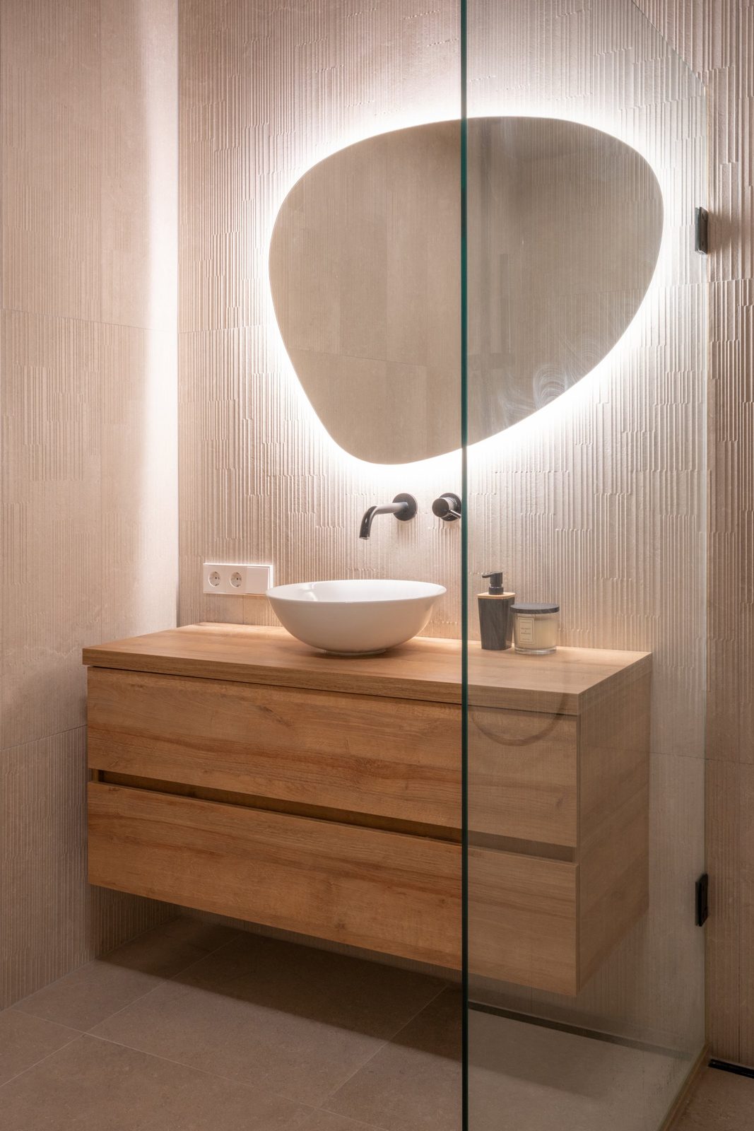 Bathroom with a backlit organic mirror, oak vanity and white vessel basin on textured tiles