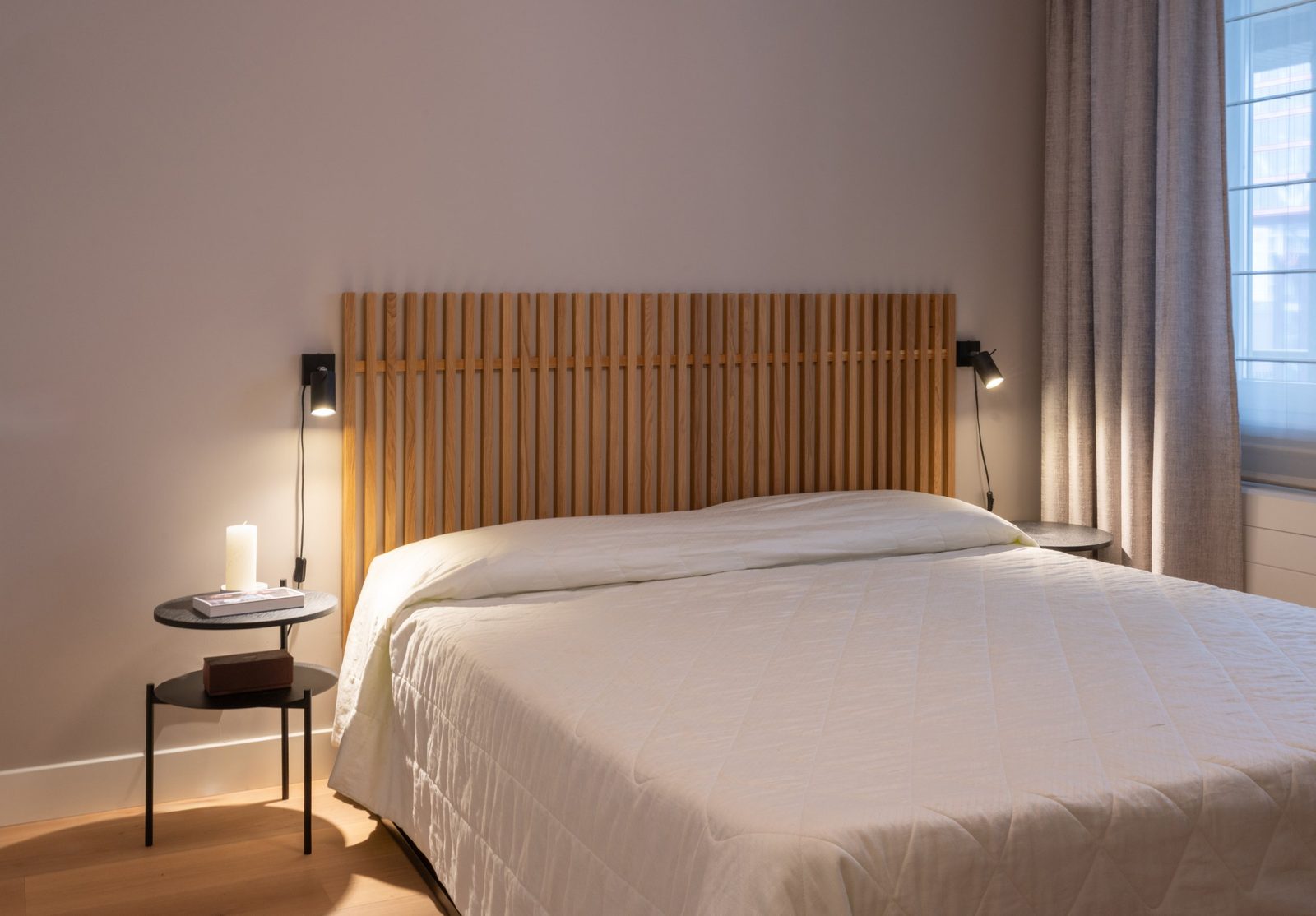 Slatted oak headboard with black wall lights, round bedside table and linen curtains
