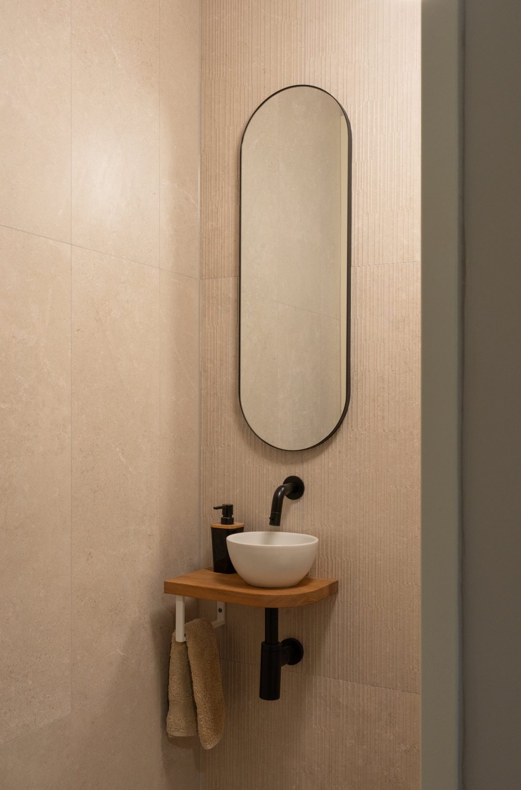 Oval black-framed mirror above a small basin on an oak shelf in a beige-tiled guest toilet