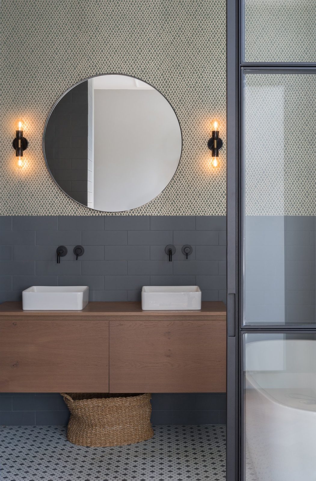 Double oak vanity with white basins, round mirror and patterned wallpaper above grey tiles