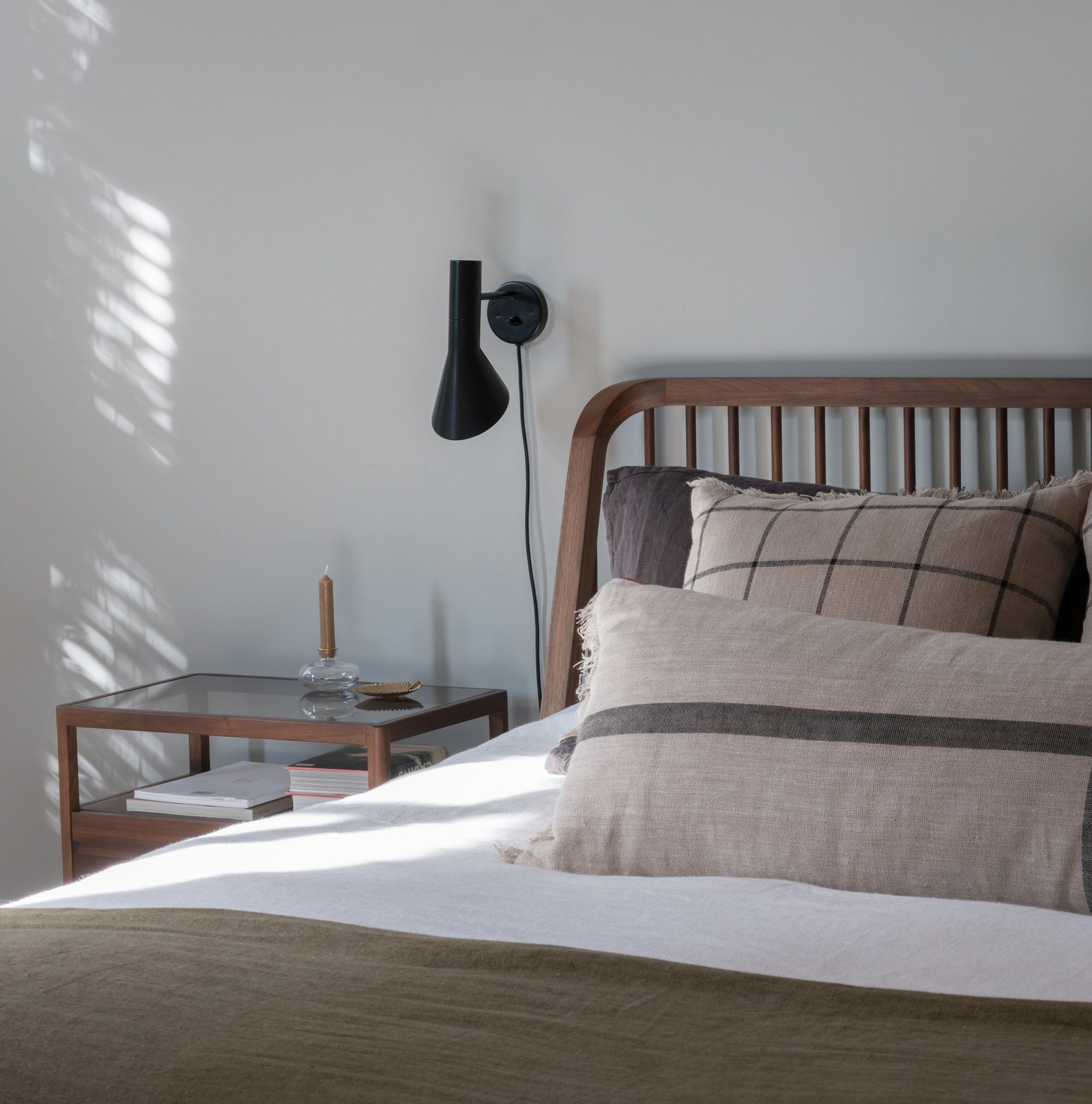 Walnut spindle bed with linen cushions, black wall light and glass-topped bedside table