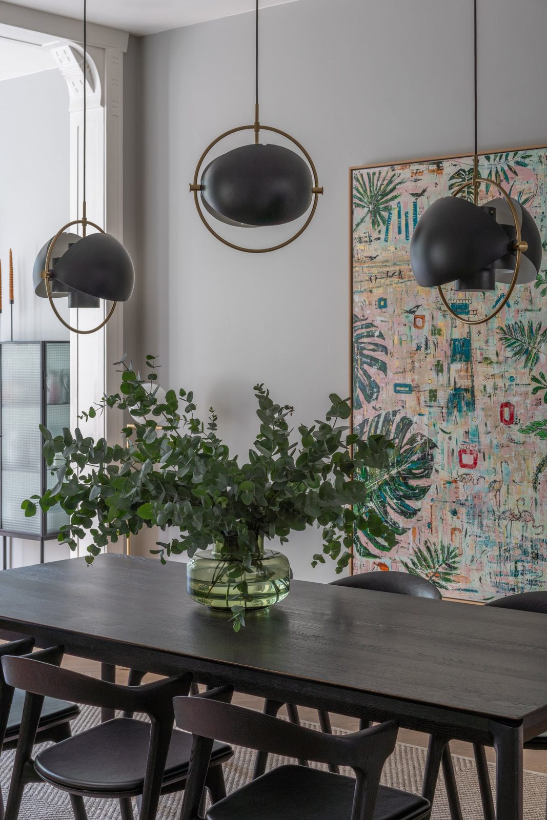 Black oak dining table under three black and brass pendants, with a large colourful painting