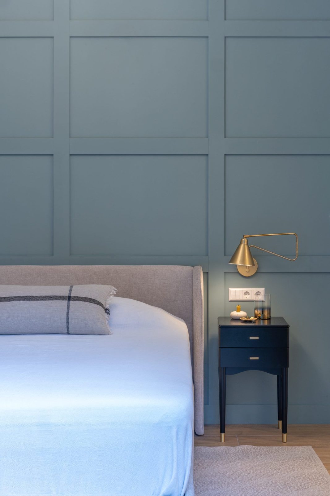 Upholstered headboard, dark bedside table and brass wall light on panelling