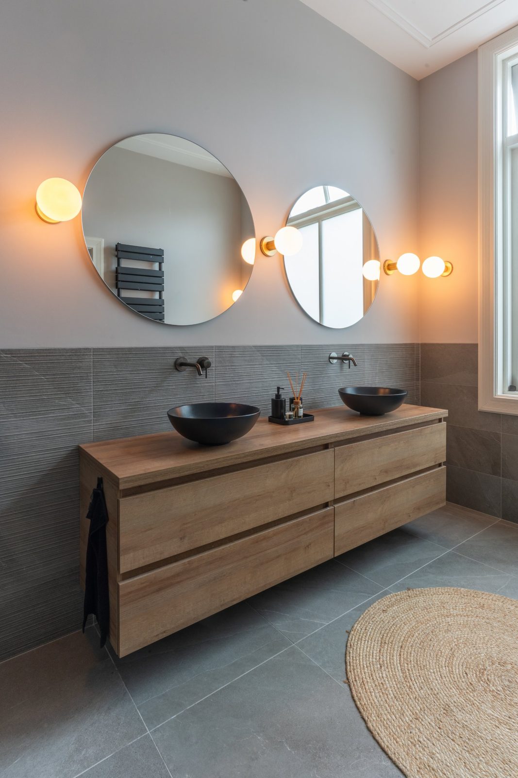 Double oak vanity with black basins and two round mirrors in a grey-tiled bathroom
