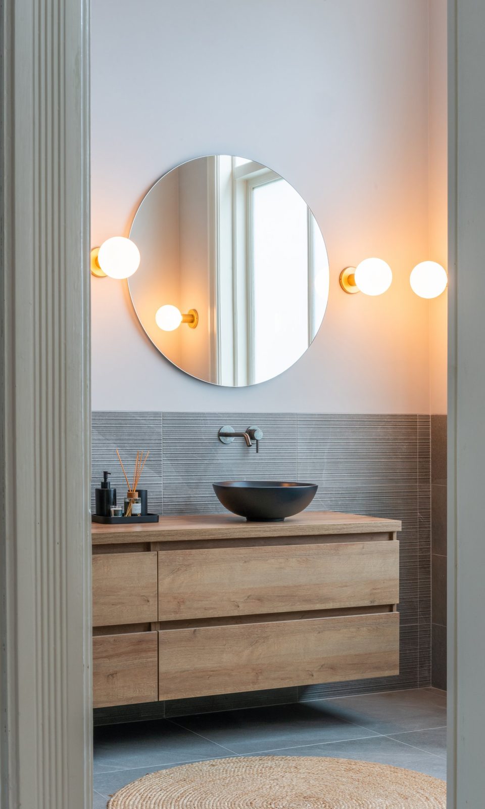 Bathroom with an oak vanity, black vessel basin, round mirror and globe wall lights
