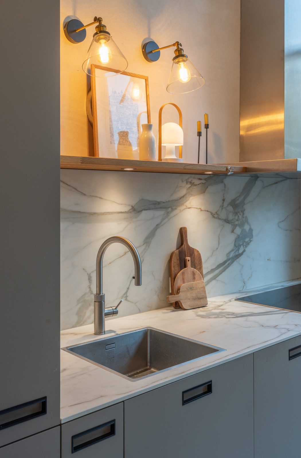 Stainless steel sink, marble splashback and oak shelf lit by brass wall lights