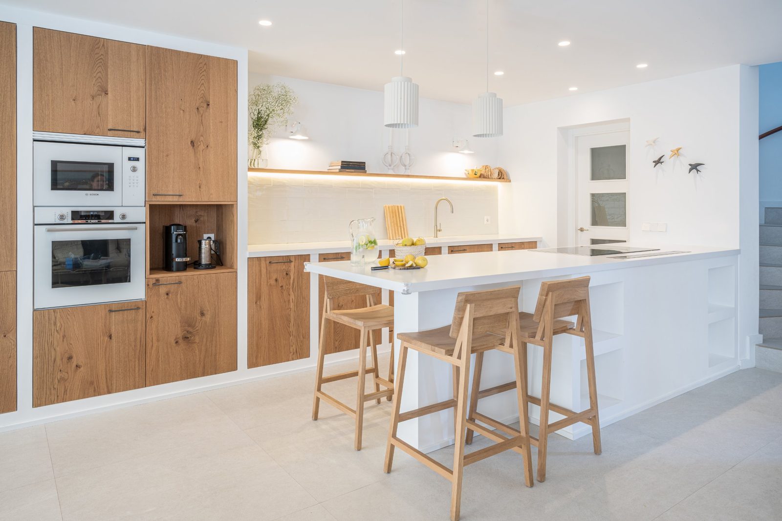 White kitchen island with oak bar stools, oak cabinetry and a lit open shelf
